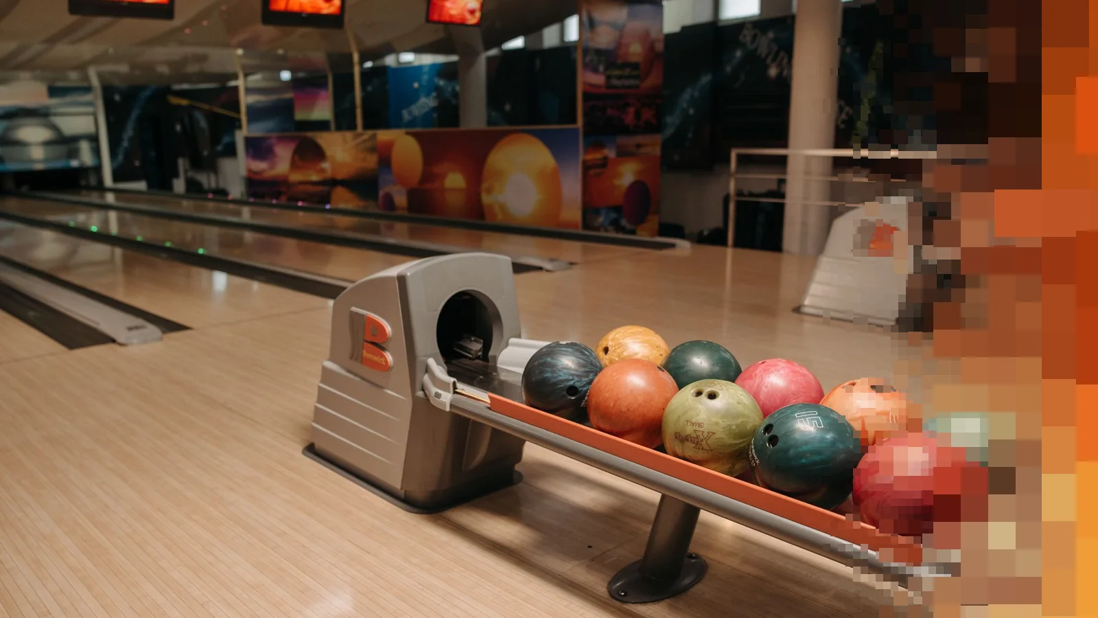 Fargerike bowlingkuler i et kulestativ i en lys bowlinghall med baner i bakgrunnen