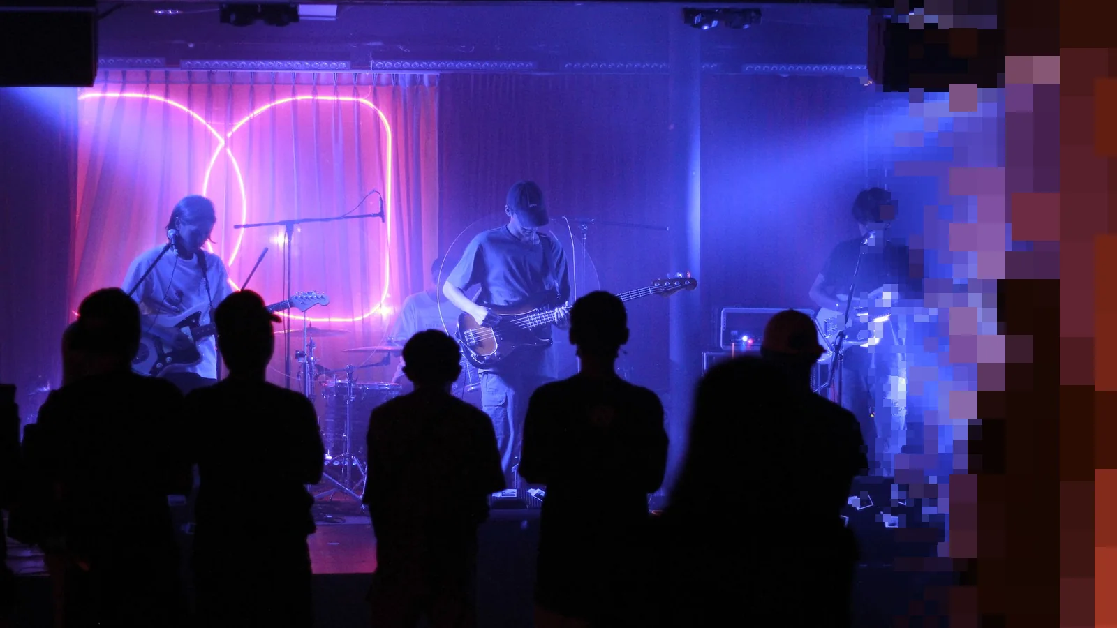 Et band spiller på en liten klubbscene i blått og lilla lys, med publikum i silhuett foran scenekanten