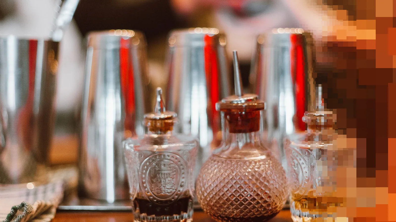 Tre små krystallflasker med bitter på en bardisk av tre, med blanke shakere og en uskarp bartender i bakgrunnen