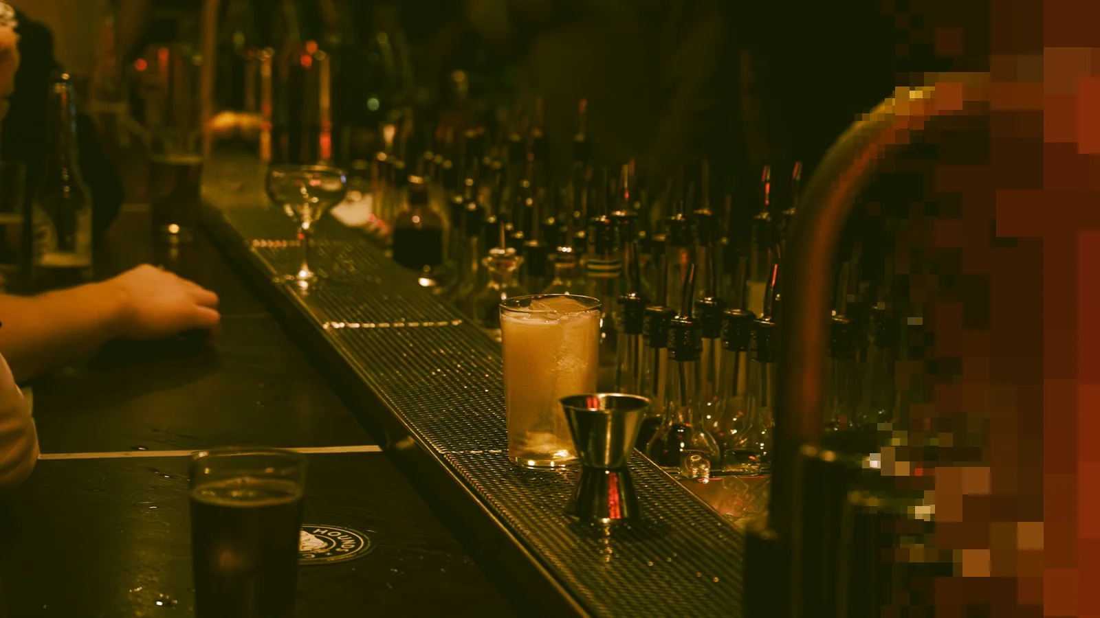 En mørk bardisk med en jigger og en lys drink foran rader med flasker og kraner, i varmt, dempet lys