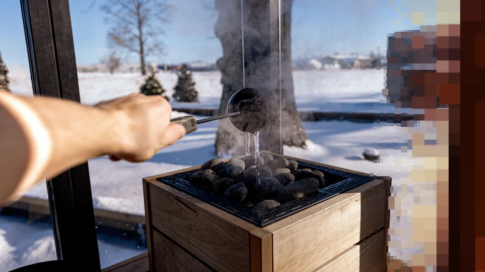 En hånd heller vann med en øse over varme steiner i en badstue med vindu ut mot snø og trær