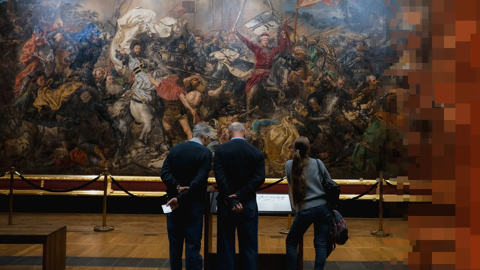 Tre besøkende sett bakfra foran et enormt historisk slagmaleri i et museum, bak et lavt avsperringstau