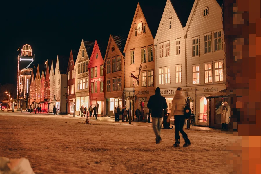 Bryggen i Bergen en vinterkveld, med opplyste trehus og folk på vei gjennom snøen