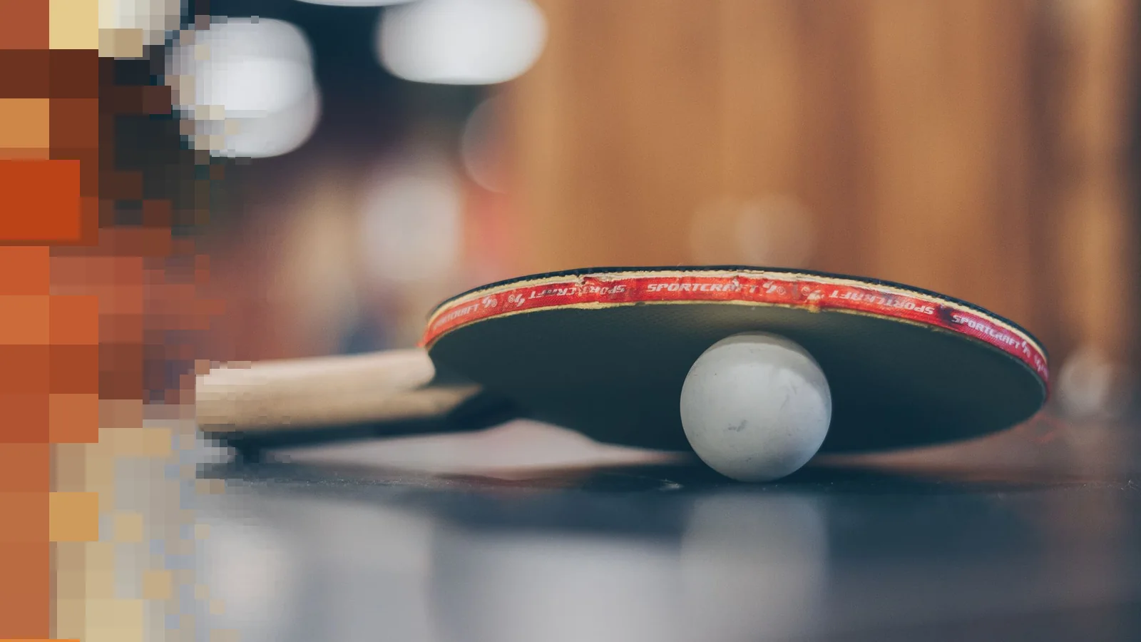 Et bordtennisracket og en hvit ball på et mørkt bord, med uskarp bakgrunn i varmt lys