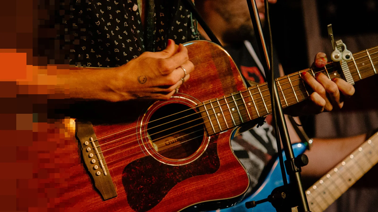 Nærbilde av hender som spiller på en akustisk gitar foran et mikrofonstativ, med en elgitar i bakgrunnen