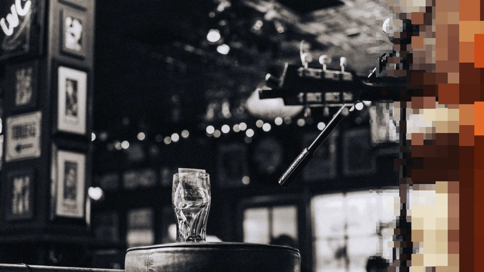 Svart-hvitt bilde fra en pub med et tomt ølglass på en barkrakk foran et mikrofonstativ og en gitar, med lysslynger i taket