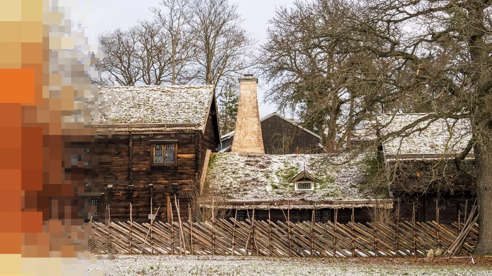Gamle tømmerhus med torvtak og et gjerde av staur, med et tynt lag snø på bakken