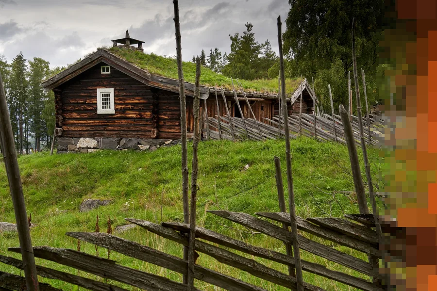 Gammelt tømmerhus med torvtak og et gjerde av staur i et grønt friluftsmuseum