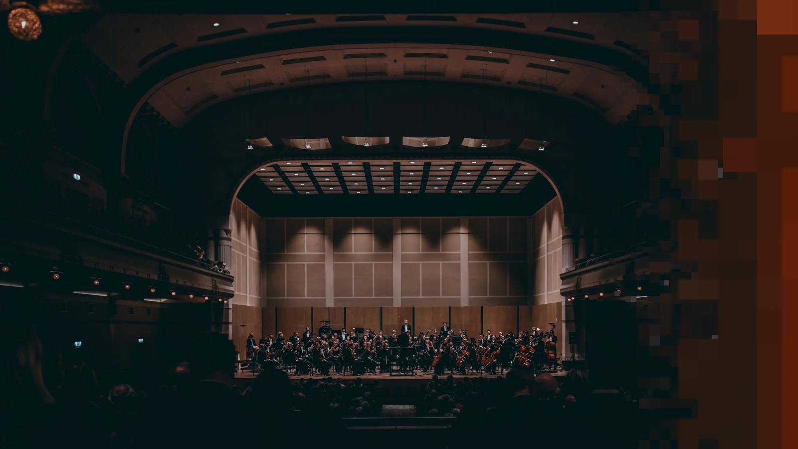 Et symfoniorkester på en opplyst scene i en stor, mørk konsertsal sett fra bakerste del av salen