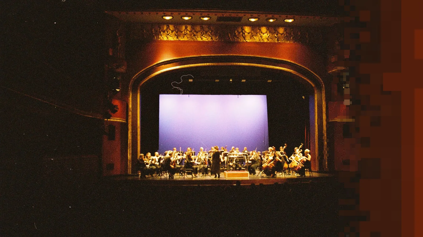 Orkester på en opplyst scene i en stor konsertsal sett fra salen