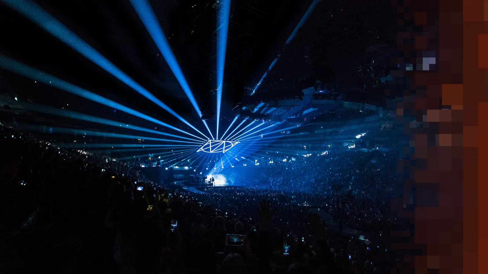 Blå lysstråler fra taket i en stor mørk konsertarena fylt med publikum