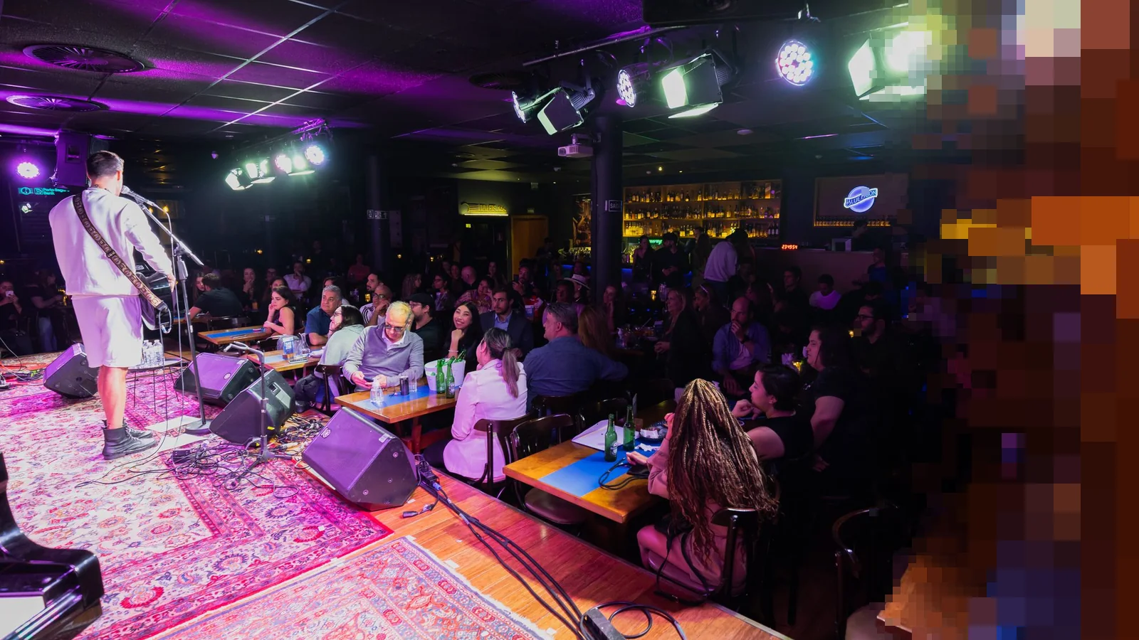 Konsertsal i lilla og blått lys med en musiker på en liten scene og publikum ved bord