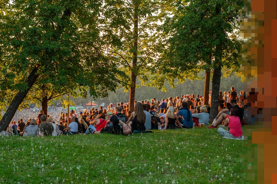 Publikum sitter tett i gresset under store løvtrær i en park en sommerkveld