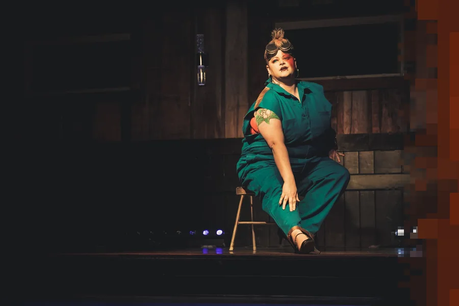 En artist i grønn kjeledress sitter på en krakk på en mørk teaterscene