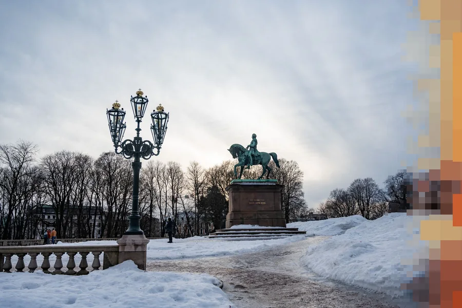 Snødekt park i Oslo med rytterstatue og en pyntet gatelykt mot en overskyet vinterhimmel