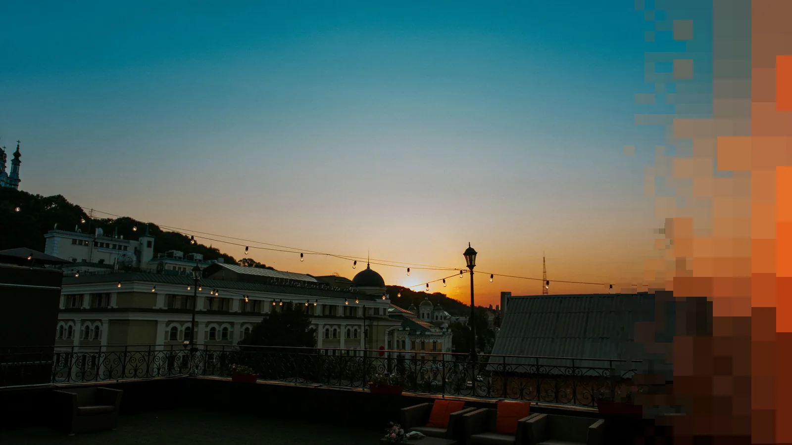 En tom takterrasse med loungesofaer og lyslenker over byens tak i solnedgang, med oransje himmel bak kupler og skorsteiner