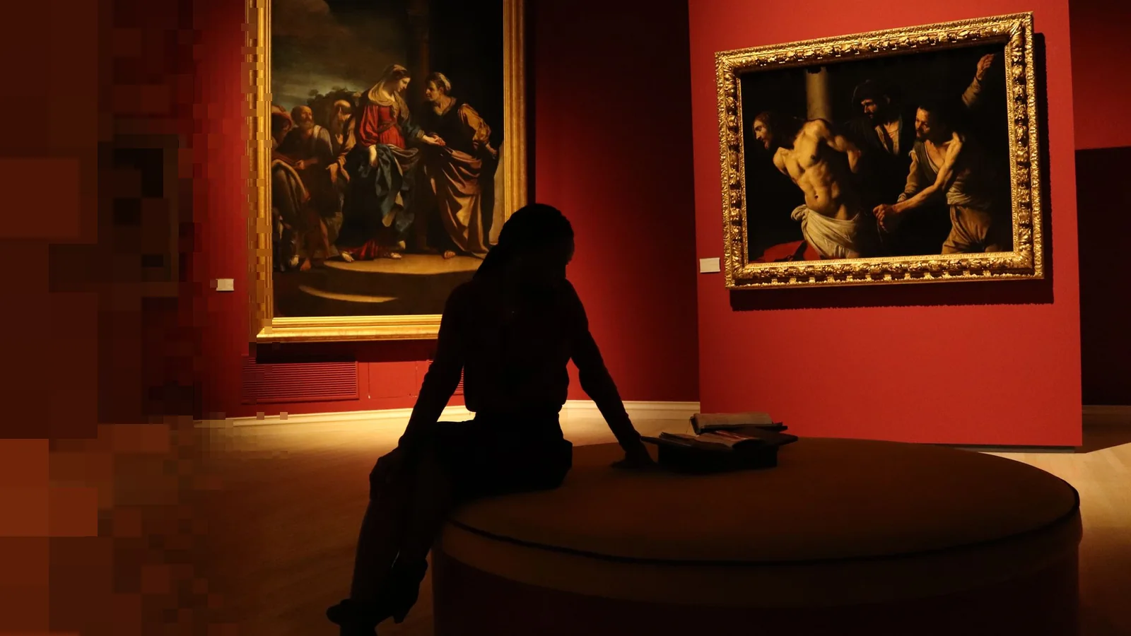 Silhuetten av en person som sitter på en rund benk i et museumsrom med røde vegger og gamle malerier