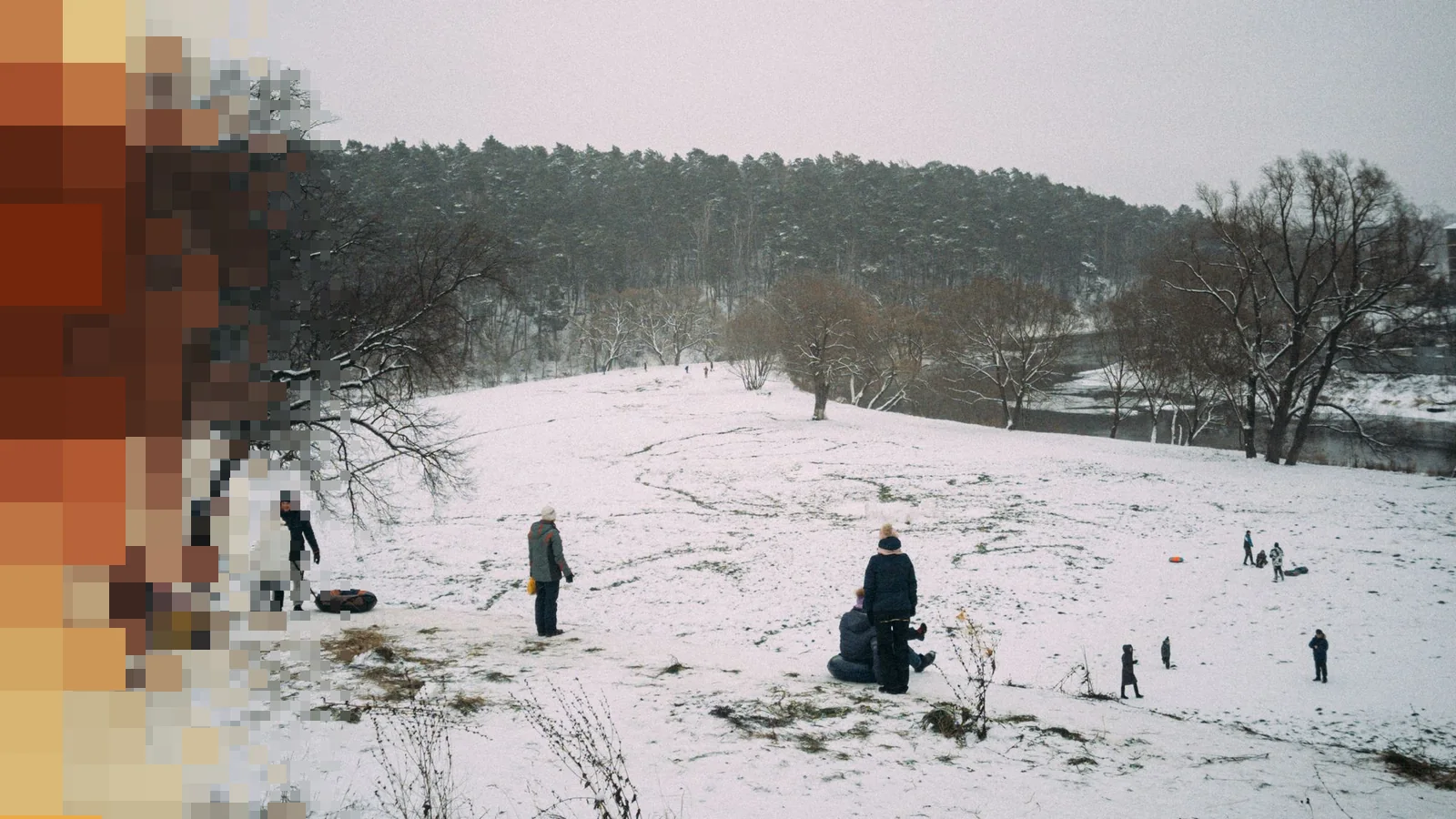 Familier med kjelker og akebrett på en snødekt bakke under en grå himmel, med nakne trær og skog i bakgrunnen